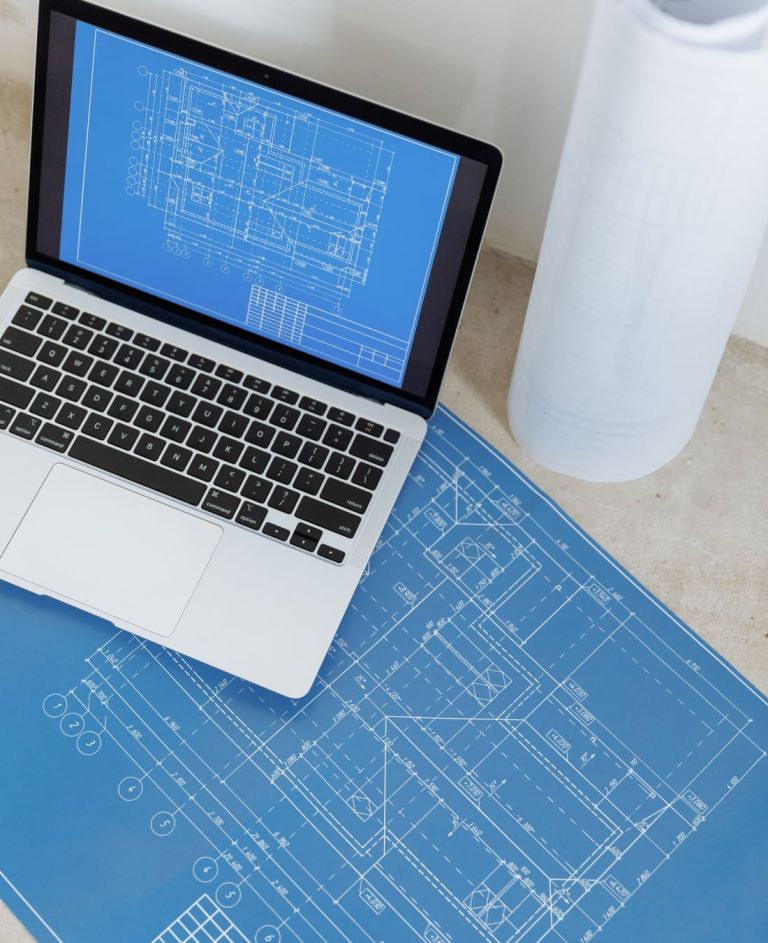 Laptop mit einer Bauzeichnung auf dem Bildschirm, darunter ein ausgebreiteter Plan, dahinter gerollte Pläne